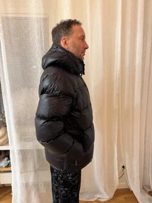 Svart pufferjacka med huva - Säljer en svart pufferjacka med stor, mysig huva och breda kanaler. Jackan är vadderad och har en glansig finish som ger en cool look. Perfekt för kalla dagar när du vill hålla värmen och ändå se snygg ut.