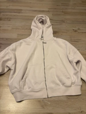 Vit hoodie med dragkedja - Säljer en vit hoodie med dragkedja framtill och två stora fickor. Hoodien har en klassisk huva och ribbade muddar vid ärmslut och nederkant. Perfekt för en avslappnad och stilren look.