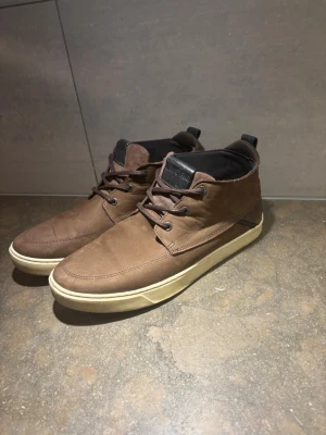 Bruna stövletter i mocka-look - Snygga bruna stövletter med mocka-liknande yta och vita gummisulor. Skorna har snörning framtill och svart innerfoder. Perfekta för dig som vill ha en stilren och avslappnad look.