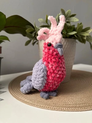 Virkad papegoja i rosa och grått - Handgjord virkad papegoja i mjukt garn med rosa kropp, ljusrosa tofs på huvudet och grå vingar, näbb och fötter. Fylld med stoppning och har bruna säkertsögon. Perfekt som dekoration eller gosedjur.
