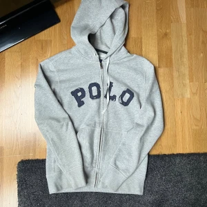 Grå hoodie med dragkedja från Polo - Snygg grå hoodie från Polo med dragkedja framtill och stor huva. Klassisk logga i mörkblått på bröstet och två fickor framtill. Perfekt för en avslappnad och stilren look. Tillverkad i mjukt bomullsmaterial🙌✅