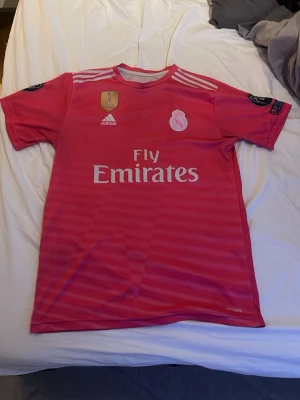 Real Madrid Ronaldo matchtröja rosa - Säljer en rosa Real Madrid matchtröja från Adidas med Ronaldo 7 på ryggen. Tröjan har vita detaljer, klubbmärke och Fly Emirates-tryck på bröstet samt Champions League-märken på ärmarna. Tillverkad i lätt och ventilerande material, perfekt för fotbollsträning.