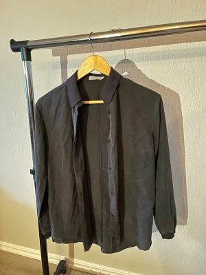 Svart skjorta från J.Lindeberg - Stilren svart skjorta från J.Lindeberg med klassisk krage och knappar framtill. Skjortan har lång ärm och är tillverkad i ett tunt, mjukt material som ger en clean look. Perfekt för dig som gillar minimalistisk stil.