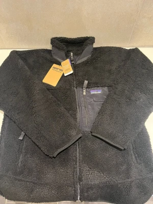 Svart teddyjacka från Patagonia - Mysig svart teddyjacka från Patagonia med hög krage och dragkedja framtill. Jackan har en bröstficka med dragkedja och detaljer i nylon, samt två sidofickor med dragkedja. Perfekt för kalla dagar och riktigt skön att bära.