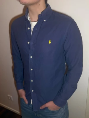 Blå skjorta från Ralph Lauren - Snygg marinblå skjorta från Ralph Lauren med klassisk button-down krage och gul broderad logga på bröstet. Skjortan har långa ärmar, vita knappar och är tillverkad i bomull. Perfekt för en clean och stilren look.