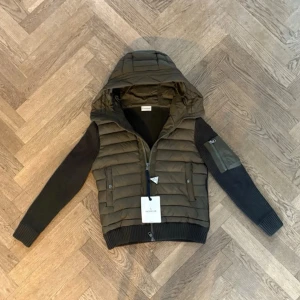 Moncler grön pufferjacka med huva - Snygg grön pufferjacka från Moncler med stickade svarta ärmar och huva. Jackan har dragkedja framtill, två fickor med dragkedja och en extra ficka på ena ärmen. Perfekt för kalla dagar och riktigt trendig look.