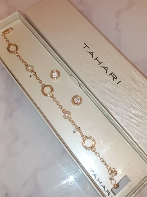 Guldfärgat armband med pärlor Tahari - Snyggt armband från Tahari i guldfärgad metall med runda länkar och vita pärlor. Armbandet har en modern design med cirkelformade detaljer och små gnistrande stenar. Perfekt accessoar för att lyfta din outfit.