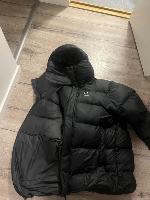 Svart pufferjacka från Haglöfs - Säljer en svart pufferjacka från Haglöfs med stor huva och quiltad design. Jackan har dragkedja framtill och är tillverkad i ett glansigt, tåligt material som håller värmen under kalla dagar. Perfekt för dig som vill ha en riktigt varm och stilren vinterjacka.