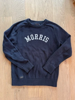 Mörkblå stickad tröja från Morris - Säljer en mörkblå stickad tröja från Morris med vit text framtill. Tröjan har rund halsringning, ribbade muddar och är långärmad. Perfekt för dig som gillar klassisk och stilren design.