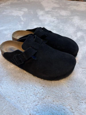 Svarta Birkenstock Boston tofflor - Säljer ett par svarta Birkenstock Boston tofflor i mocka med justerbart spänne och klassisk korksula. Perfekta för dig som vill ha både stil och komfort. Passar till chill vardagsstil och är riktigt sköna att glida runt i hemma.