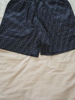 Svarta Fendi shorts med logomönster - Snygga svarta shorts från Fendi med heltryckt logomönster i mörkgrått. Shortsen har elastisk midja med snörning och är perfekta för en trendig och avslappnad stil. Materialet känns lätt och bekvämt, perfekt för varma dagar.