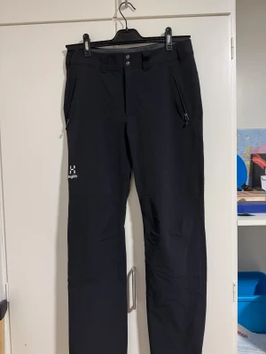 Svarta täckbyxor från Haglöfs - Svarta täckbyxor från Haglöfs med snygga silverfärgade knappar och dragkedjor på fickorna. Byxorna har en rak passform och är gjorda i slitstarkt syntetmaterial, perfekta för utomhusaktiviteter. Broderad Haglöfs-logga på benet ger en clean look. Aldrig använt de eftersom att de är stora på mig.