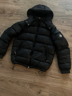 Moncler vezere - Snygg svart dunjacka från Moncler med huva och dragkedja framtill. Jackan har två snedställda fickor med dragkedja och en Moncler-logga på ärmen. Puffig modell med justerbar nederkant och ribbade muddar vid ärmslut. Perfekt för kalla vinterdagar.