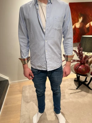 Ljusblå skjorta med rutiga detaljer - Snygg ljusblå skjorta med klassisk krage och långa ärmar. Skjortan har rutigt tyg på insidan av manschetterna som syns när du viker upp ärmarna. Perfekt för en avslappnad men stilren look.