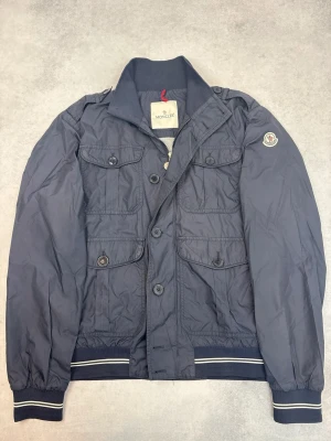 Mörkblå vindjacka från Moncler - Snygg mörkblå vindjacka från Moncler med fyra framfickor, ribbade muddar och krage. Jackan har Moncler-logga på ärmen och stängs med knappar. Perfekt för dig som vill ha en stilren och sportig look. Materialet är lätt och vindtätt.
