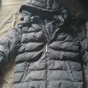 Svart pufferjacka från GANT - Snygg mörkblå pufferjacka från GANT med huva och dragkedja framtill. Jackan har quiltad design, två fickor med dragkedja och diskret logga på bröstet. Perfekt för kalla dagar. Skicka dm för frågor. Pris ej hugget i sten 
