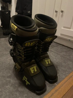 Full tilt pjäxor  - Säljer ett par FT BE skistövlar från Full Tilt i svart och olivgrön med gula detaljer och spännen. Stövlarna har tydliga FT-loggor och BE-märkning, samt flera justerbara spännen för bra passform. Perfekta för dig som gillar freestyle eller parkåkning.