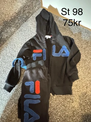 Svarta mjukisbyxor från FILA - Säljer ett par svarta mjukisbyxor från FILA med stora blå och röda loggor längs benet. Byxorna har resår i midjan och muddar vid bensluten för skön passform. Perfekta för chill dagar eller när du vill ha det bekvämt.