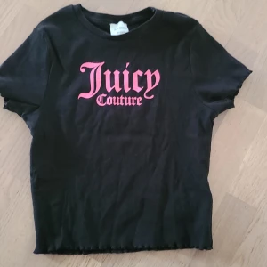 Juicy Couture t-shirt med rosa tryck - Cool svart t-shirt från Juicy Couture med stort rosa logotryck på bröstet. Modellen är kortärmad och har en ribbad struktur i tyget samt vågig kant nedtill och vid ärmslut. Perfekt för dig som gillar statement-plagg och streetstyle.