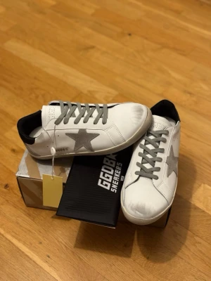 Golden Goose vita  - Snygga vita sneakers från Golden Goose med grå stjärna på sidan och grå skosnören. Skorna har svarta detaljer vid hälen och en klassisk låg siluett. Tillverkade i läder med slitna detaljer för en cool, avslappnad look.