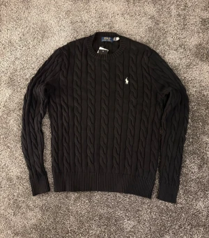 Svart kabelstickad tröja Polo Ralph Lauren - Kabelstickad svart tröja från Polo Ralph Lauren med rund halsringning och den klassiska broderade loggan i vitt på bröstet. Tröjan har ribbade muddar vid ärmslut och nederkant, perfekt för en stilren och tidlös look.