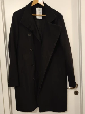 Svart kappa från Emilio Denim Issue - Stilren svart kappa från Emilio Denim Issue med klassisk krage och knäppning framtill. Ca 102cm lång. Knappt använd. Storlek saknas men bör vara L, bystmått: 124cm (62cm från armhåla till armhåla)