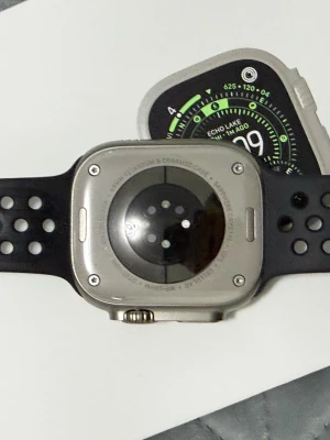 Apple Watch Ultra 3  med svart sportarmband - Apple Watch Ultra med robust boett i titan och keramisk baksida. Klockan har en fyrkantig form och svart sportarmband med hål för bättre ventilation. Skärmen är stor och tydlig, perfekt för träning och vardag. Snygg och modern design.