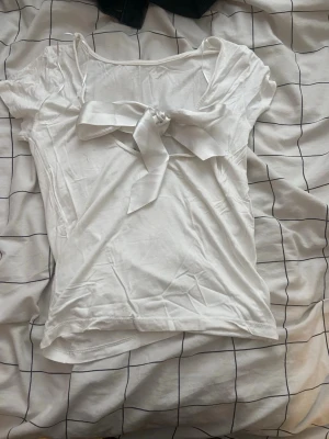 Vit topp med rosett och kort ärm - Söt vit topp med korta ärmar och en stor rosett framtill vid urringningen. T-shirten har en feminin och enkel design, perfekt att styla med jeans eller kjol. Tillverkad i mjukt bomullsmaterial för en bekväm känsla.