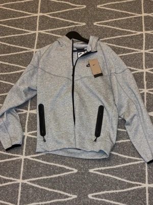 Grå hoodie med dragkedja från Nike - Snygg gråmelerad hoodie från Nike med hel dragkedja och svarta detaljer på fickorna. Hoodien har huva, långa ärmar och klassisk Nike-logga på bröstet. Perfekt för en sportig och avslappnad stil.