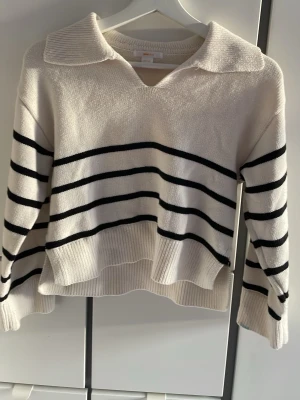 Randig stickad tröja från Gina Tricot - Säljer en vit stickad tröja från Gina Tricot med svarta horisontella ränder. Tröjan har en bred krage, v-ringning och långa ärmar. Modellen är lite loose och har ribbade muddar vid ärmslut och nederkant.