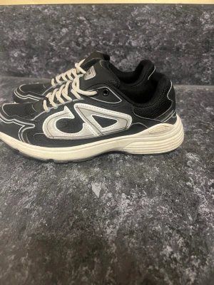 Svarta Dior sneakers med vita detaljer - Säljer ett par svarta Dior sneakers med vita detaljer och vita snören. Skorna har en chunky sula och meshpaneler för extra komfort. Snygg kontrast mellan svart och vitt samt tydlig Dior-logga på sidan. Perfekta för dig som gillar streetwear och exklusiva märken.