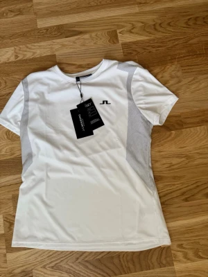 Vit sport-t-shirt från J.Lindeberg - Vit sport-t-shirt från J.Lindeberg med grå meshpaneler på sidorna. Tillverkad i TX Jersey-material med 4-vägsstretch, snabbtorkande och fukttransporterande egenskaper. Perfekt för träning eller sportaktiviteter. Diskret logga på bröstet.