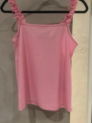 Rosa linnetopp med volangband - Söt rosa linnetopp med breda volangprydda axelband. Toppen har en rak passform och är tillverkad i ett mjukt, stretchigt material som känns bekvämt mot huden. Perfekt för varma dagar och enkel att styla med jeans eller kjol.