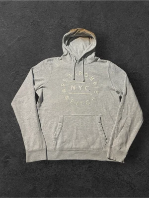 Grå hoodie från Abercrombie & Fitch | L - Snygg grå hoodie från Abercrombie & Fitch med trycket 'NYC' framtill. Hoodien har en klassisk känguruficka, dragsko i huvan och mjukt material. Perfekt för en avslappnad streetwear-look.