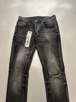 Svarta slitna jeans från Purple - Säljer ett par svarta jeans från Purple med snyggt slitna detaljer och en trasig knädel. Jeansen har klassisk femficksdesign, raka ben och tvättad finish för en cool look. Perfekta för dig som gillar streetwear och vill ha en avslappnad stil.