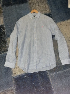 Blårutig skjorta från Ralph Lauren - Klassisk blårutig skjorta från Ralph Lauren i bomull med normal passform. Skjortan har knappar framtill, lång ärm och en liten rosa logga broderad på bröstet. Perfekt till jeans eller chinos för en clean och stilren look.