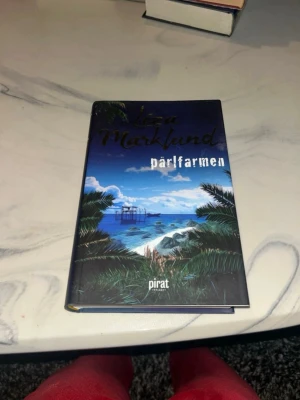 Pärlfarmen av Liza Marklund - Säljer romanen 'Pärlfarmen' av Liza Marklund, utgiven av Piratförlaget. Omslaget har en färgstark illustration med blå himmel, hav och palmer, vilket ger tropiska vibbar. Perfekt för dig som gillar spännande skönlitteratur med äventyrskänsla.