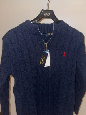 Marinblå kabelstickad tröja Polo Ralph Lauren - Kabelstickad marinblå tröja från Polo Ralph Lauren med halv dragkedja och röd broderad logga på bröstet. Tröjan har ribbade muddar och klassisk passform. Perfekt för dig som gillar stilren och tidlös design.