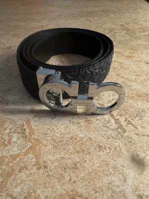 Svart bälte från Salvatore Ferragamo - Snyggt svart bälte från Salvatore Ferragamo med präglat mönster längs hela remmen och en stor, blank silverfärgad logospänne. Perfekt accessoar för att lyfta din outfit med en lyxig touch.