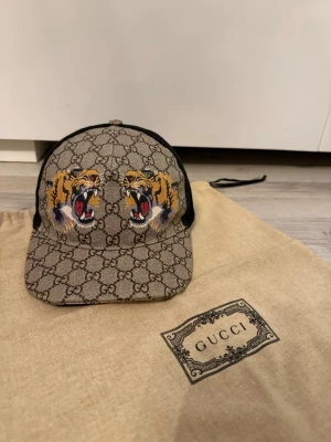Gucci keps med tigerbrodyr - Säljer en snygg Gucci keps i beige med klassiskt GG-mönster och två broderade tigrar framtill. Kepsen har svart mesh baktill och justerbart band med röd-gröna detaljer. Materialet är canvas och mesh, perfekt för dig som vill sticka ut. Dustbag följer med!!! Som ny
