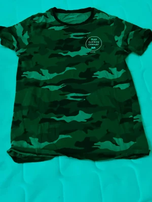 Camo t-shirt från Warp - Säljer en grön camouflagemönstrad t-shirt från Warp Clothing Company. T-shirten har rund halsringning och korta ärmar. Perfekt för dig som gillar streetstyle och vill sticka ut lite extra.