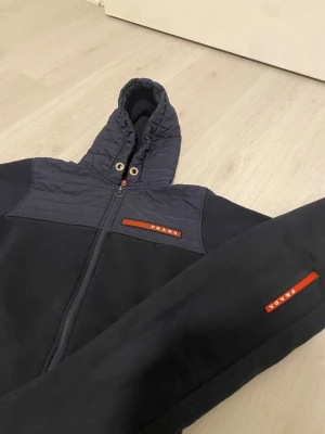 Svart hoodie från Zara med dragkedja - Snygg svart hoodie från Zara med dragkedja framtill och quiltad detalj över axlar och huva. Hoodien har två fickor, ribbade muddar och en ikonisk röd Zara -logga på bröstet. Perfekt för dig som vill ha en stilren och exklusiv look.