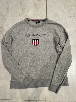 Grå tröja från GANT - Snygg grå stickad tröja från GANT som passar dig mellan ca 165-175, broderad logga och ett sköldmärke med stjärnor och ränder på bröstet. Tröjan har rund halsringning, långa ärmar och ribbade muddar