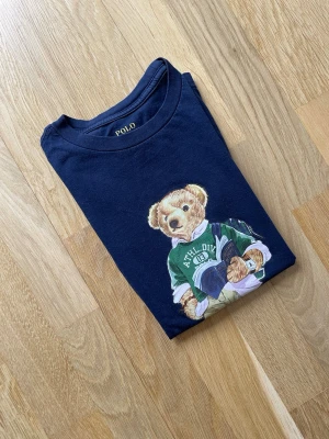 Mörkblå Polo Ralph Lauren t-shirt, strl 140 - Snygg mörkblå Polo bear t-shirt från Polo Ralph Lauren med ikoniskt björntryck på bröstet. Nypris ca 1000 kr mitt pris endast 249 kr. Fraktar inom 1 dag, om du har någon fundering så är det bara att höra av sig😊