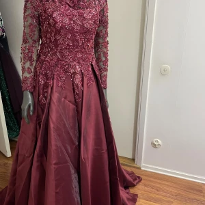 Vinröd långklänning med spets och snörning - Magisk vinröd långklänning med transparenta ärmar och vackra spetsdetaljer med broderier och pärlor. Klänningen har snörning i ryggen och en vid kjol i glansigt tyg. Perfekt för bal eller fest där du vill sticka ut.