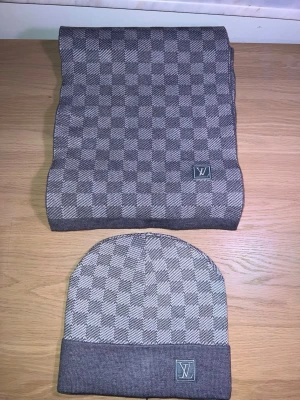 Grå rutig halsduk från Louis Vuitton - Snygg grå halsduk från Louis Vuitton med klassiskt rutigt mönster och diskret LV-logga. Halsduken och har en mjuk känsla, perfekt för att hålla värmen och addera stil till din outfit. Med 90% bomull.