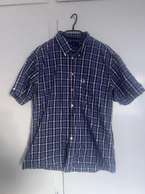 Blårutig kortärmad skjorta Fred Perry - Säljer en blårutig skjorta från Fred Perry med korta ärmar och klassisk button-down krage. Skjortan har vita knappar och en broderad logga på bröstfickan. Materialet är mjuk bomull och färgerna går i blått, vitt och rött.