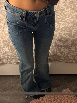 Blå bootcut jeans med låg midja - Säljer ett par blå bootcut jeans med låg midja och klassiska fem fickor från H&M i strl 36. Jeansen har råa kanter nertill och en snygg tvätt som ger en vintagekänsla. Perfekta för dig som gillar retrostil och vill ha ett par jeans som sticker ut.
