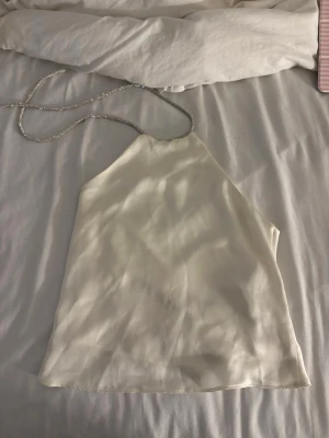 Vit siden halterneck linnetopp - Säljer en stilren vit linnetopp i siden med tunna glittriga halterneckband. Toppen har en enkel och elegant design som ger en lyxig känsla. Perfekt för dig som vill ha något trendigt och snyggt till sommaren.💞