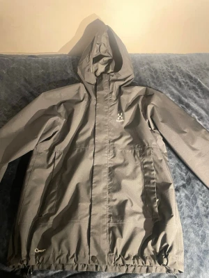 Svart vindjacka från Haglöfs - Snygg svart vindjacka från Haglöfs med huva och dragkedja framtill. Jackan har två stora fickor med dragkedja och justerbar nederkant. Perfekt för blåsiga dagar och outdoor-aktiviteter. Materialet är slitstarkt och vattenavvisande. Storleken är L men skulle rekommendera den till XL då den inte passar mig jag är 184cm. Vid frågor skicka PM. Nypris 2400kr